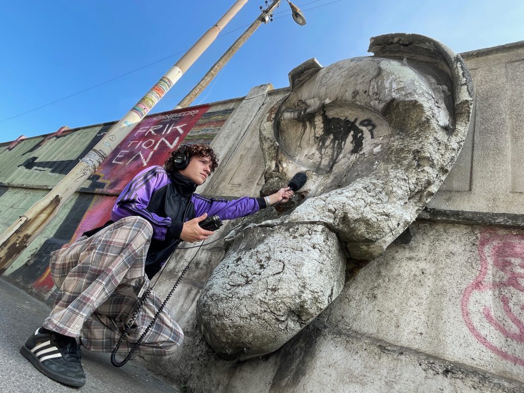 Auf dem Bild hockt ein junger Mann mit Kopfhörer und Mikrophon vor einer Betonmauer, aus der ein überdimensioniertes Ohr aus Beton herausragt. Er hält ein Mikrophon direkt in die steinerne Ohrmuschel. Im hintergrund ist an die MMauer das Wort Friktionen geschmiert.