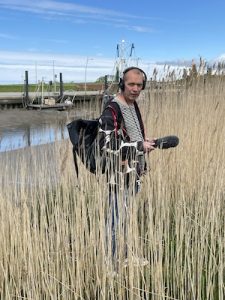 Josef M. Schäfers mit Kopfhörern und Mikrophon vor einem Fluss inmitten von Deichgras.